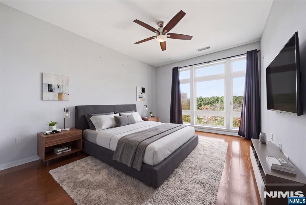 Furnished bedroom with a gray bed, hardwood floors, and large windows overlooking trees.
