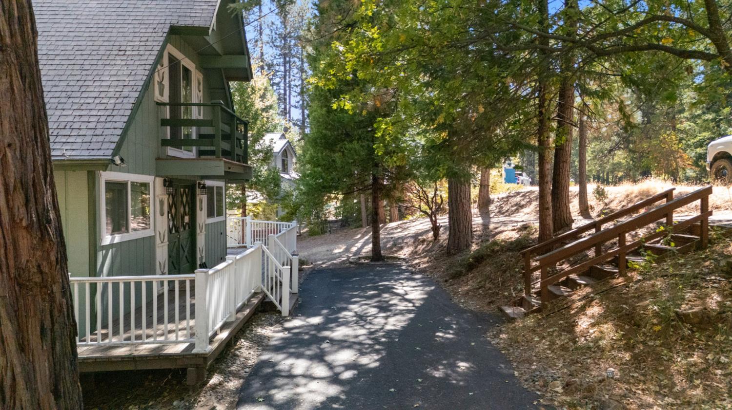 Green house with white porch railings beside a paved path and wooded hillside.