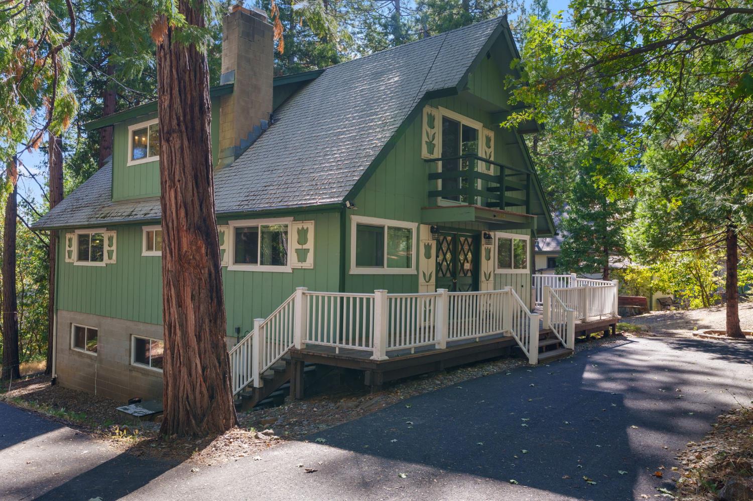 Green steep-roofed home with a raised white-railed deck among tall trees.
