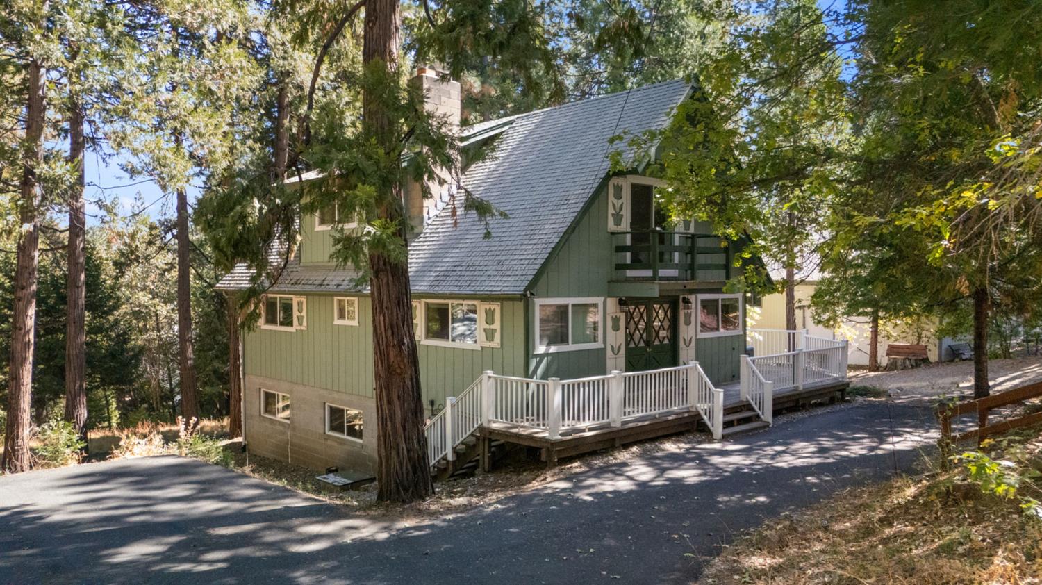 Green multi-level house with a steep roof and white-railed deck among tall trees.