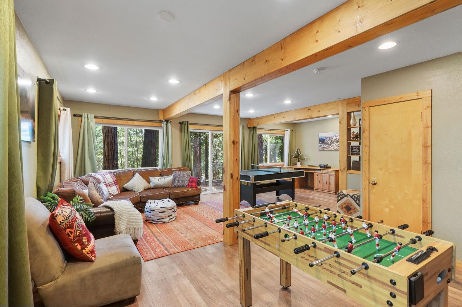 Game room with a foosball table, pool table, and sectional sofa beside large forest-facing windows.