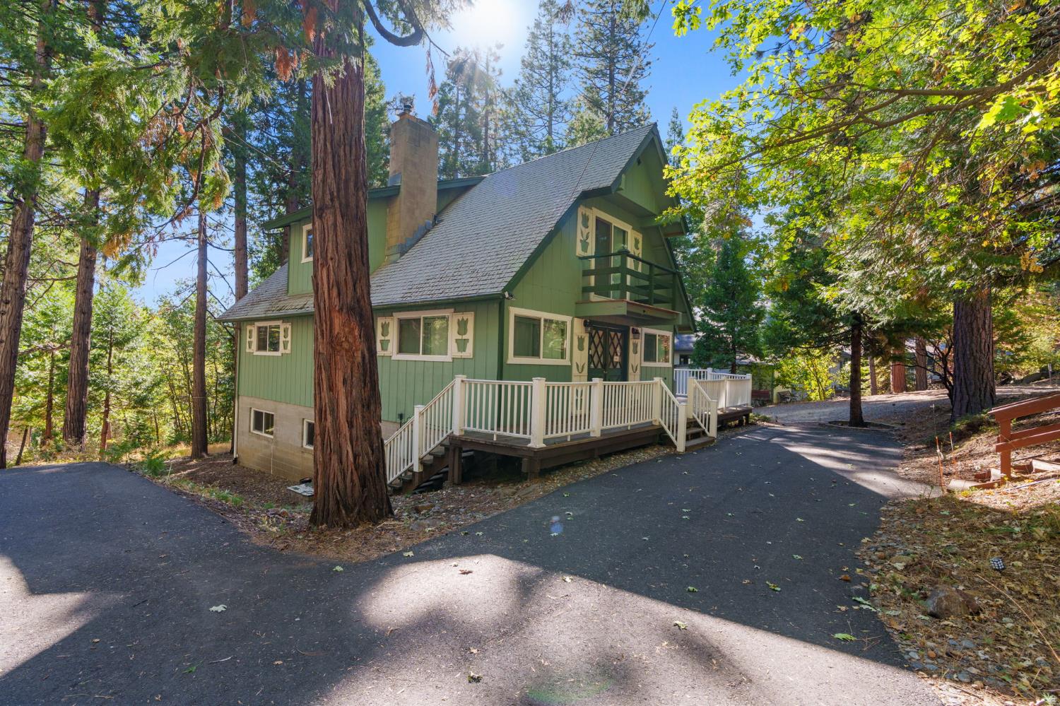 Green two-story cabin with a white-railed deck and balcony among tall trees.