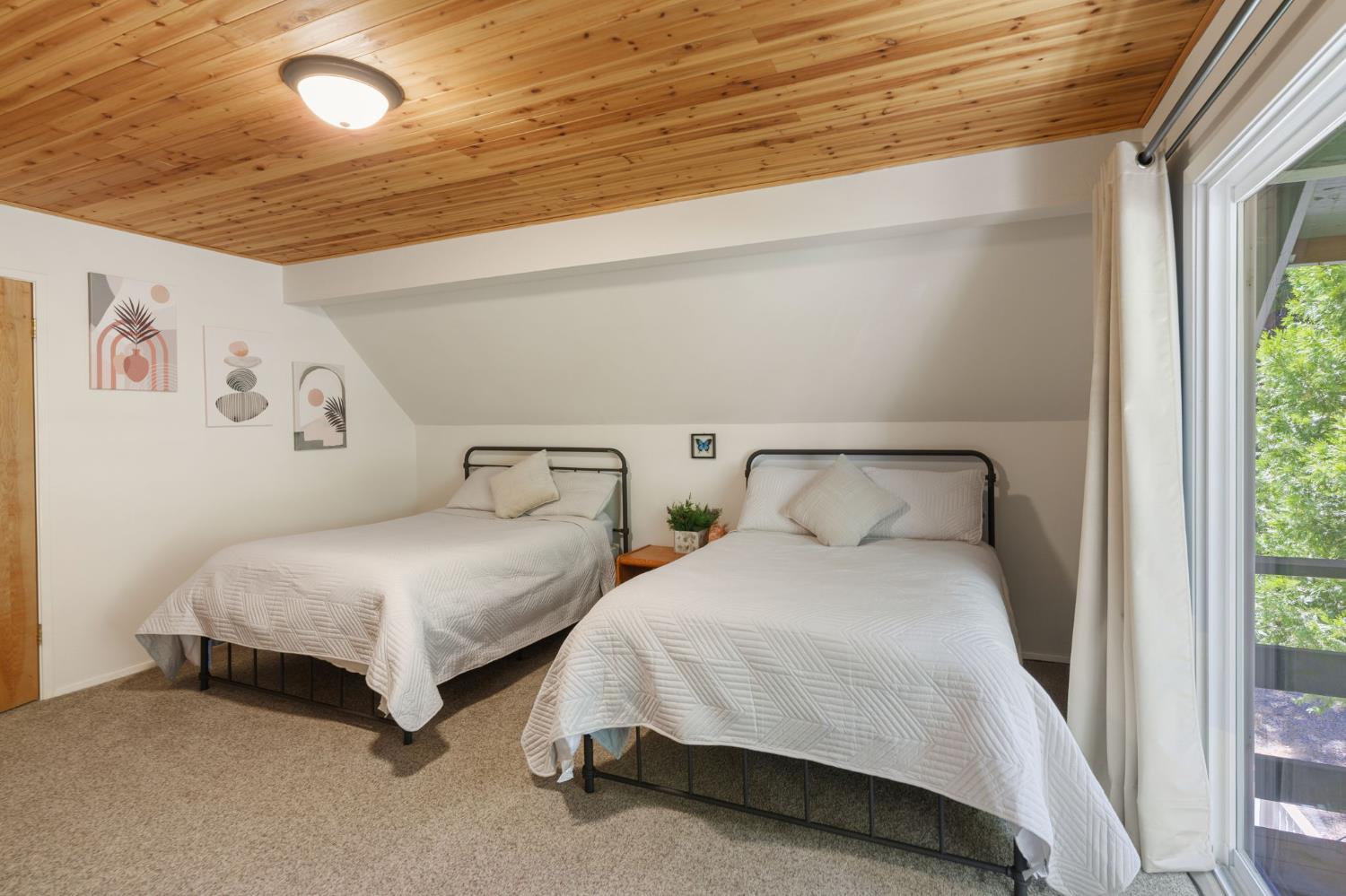 Bedroom with two beds, a sloped wood-plank ceiling, and a glass door overlooking greenery.