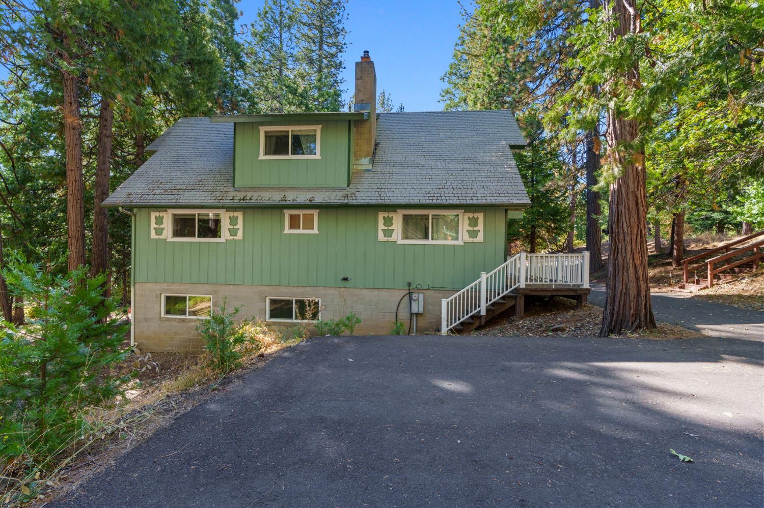 Green two-story house with a dormer, white-railed entry steps, and a paved driveway among tall pines.