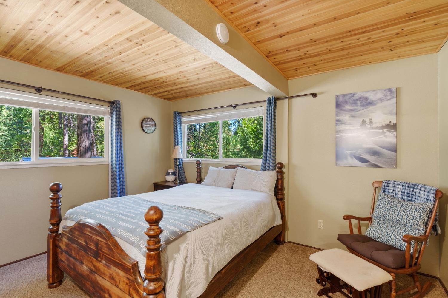 Bedroom with a wooden four-poster bed, vaulted pine ceiling, and windows overlooking trees.