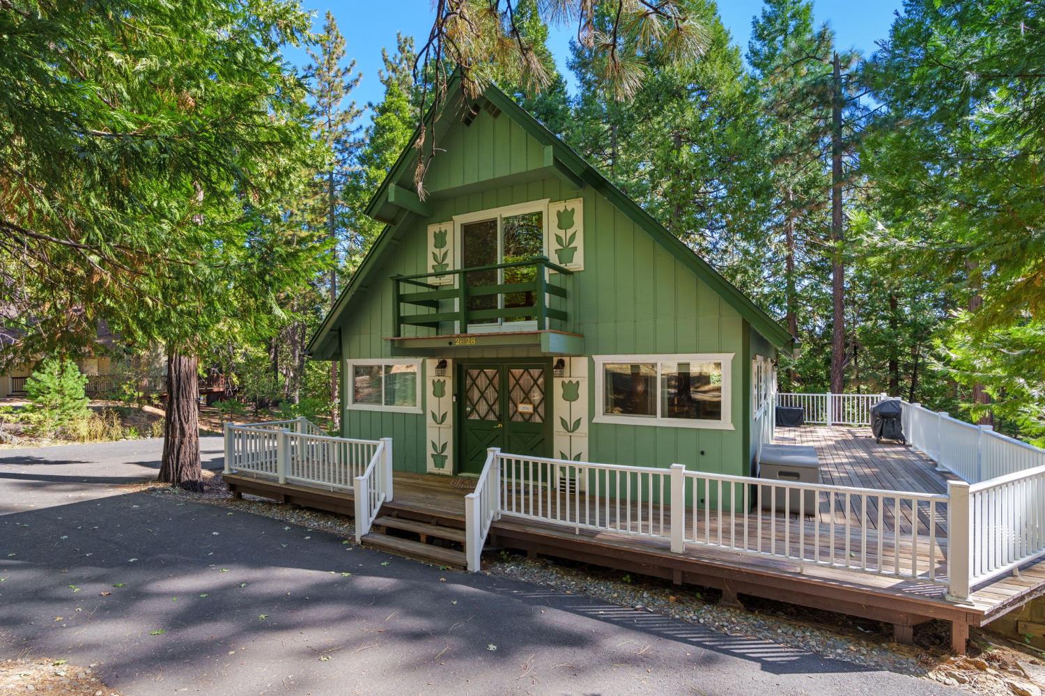 Green A-frame home with a raised white-railed deck among tall pine trees.