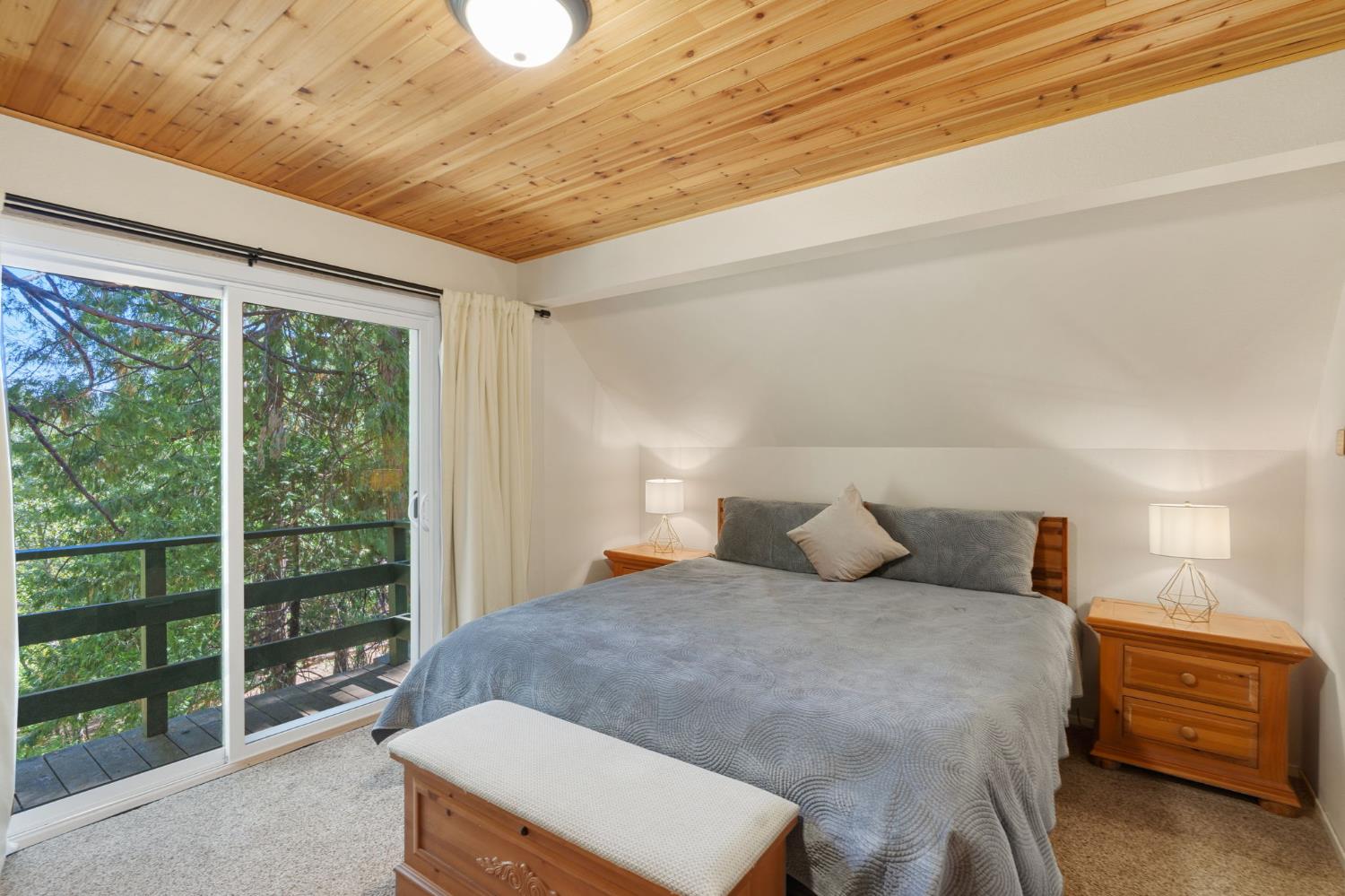 Bedroom with a wood-plank ceiling, gray bed, and sliding doors to a tree-lined balcony.