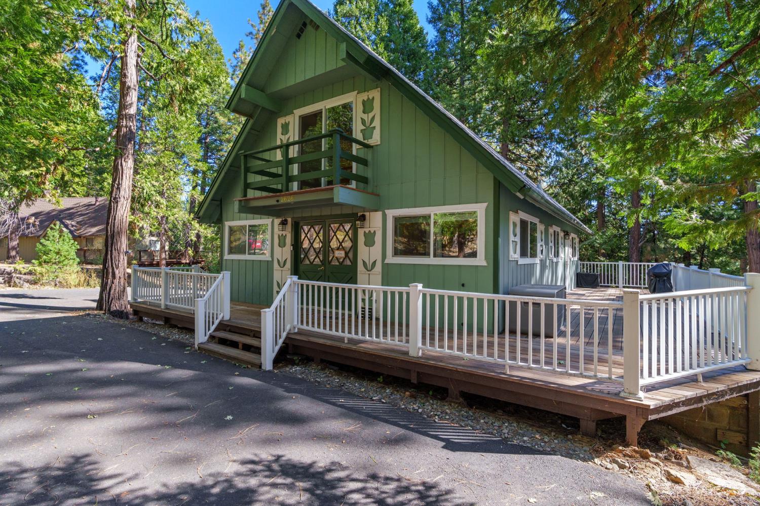 Green A-frame house with a white-railed deck nestled among tall evergreen trees.