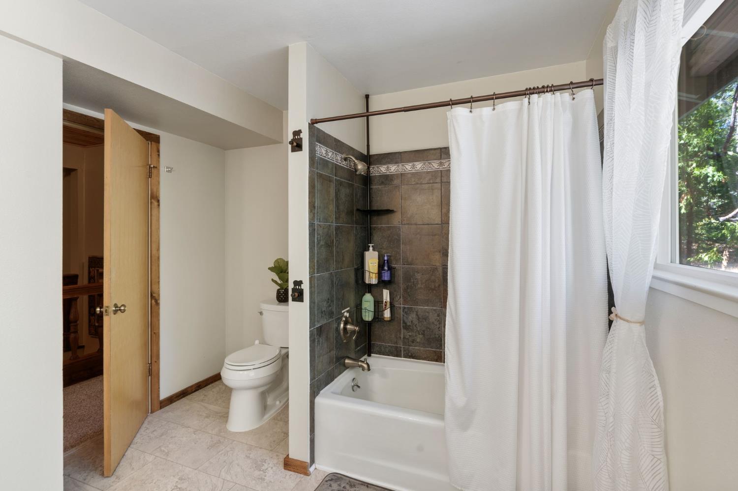 Bathroom with a dark tiled tub and shower beside a toilet and window overlooking trees.