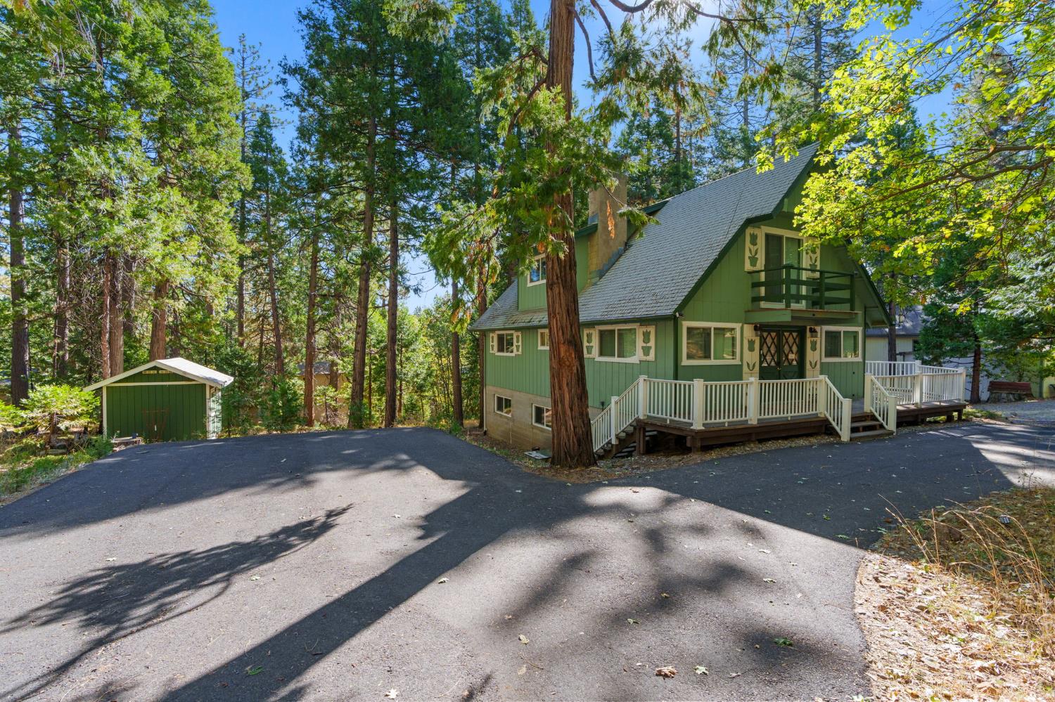 Green gabled house with a raised white-railed deck among tall trees, beside a detached shed.