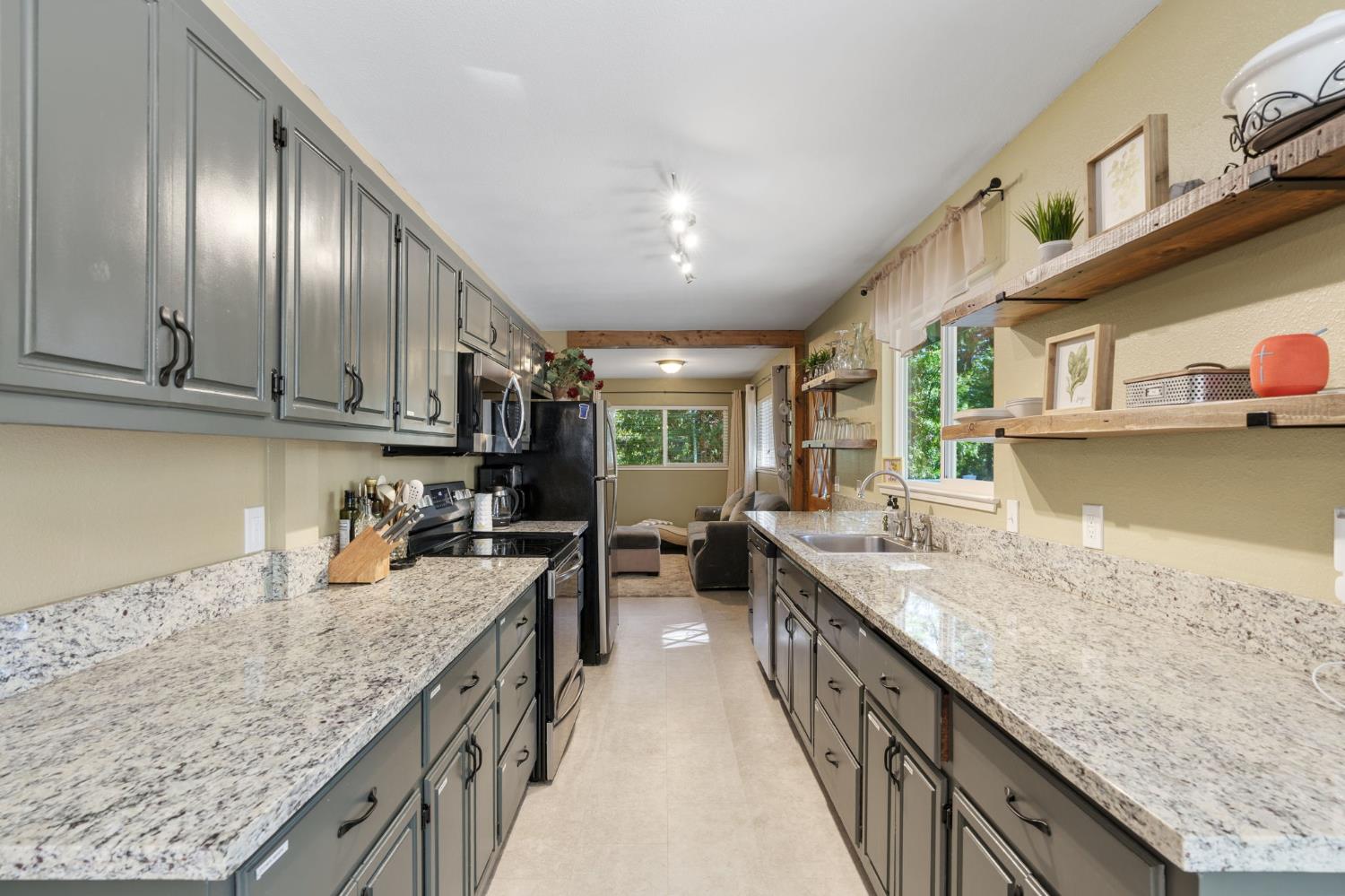 Galley kitchen with gray cabinets, granite counters, and open shelves beside large windows.