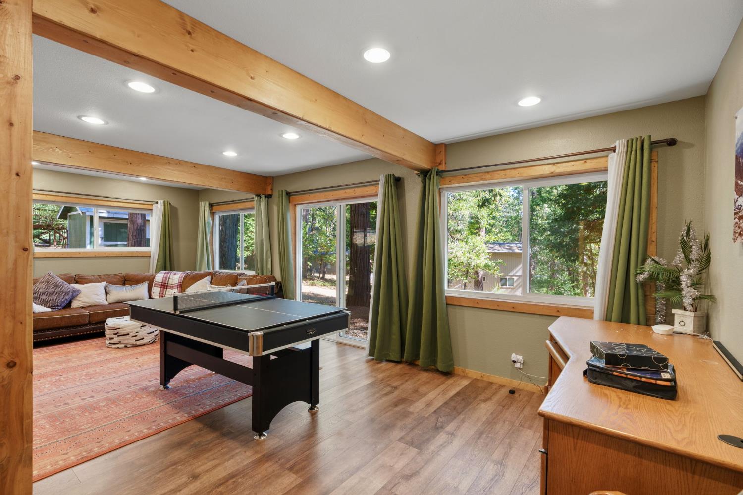 Game room with a ping-pong table, brown sofa, exposed timber beams, and large windows overlooking trees.