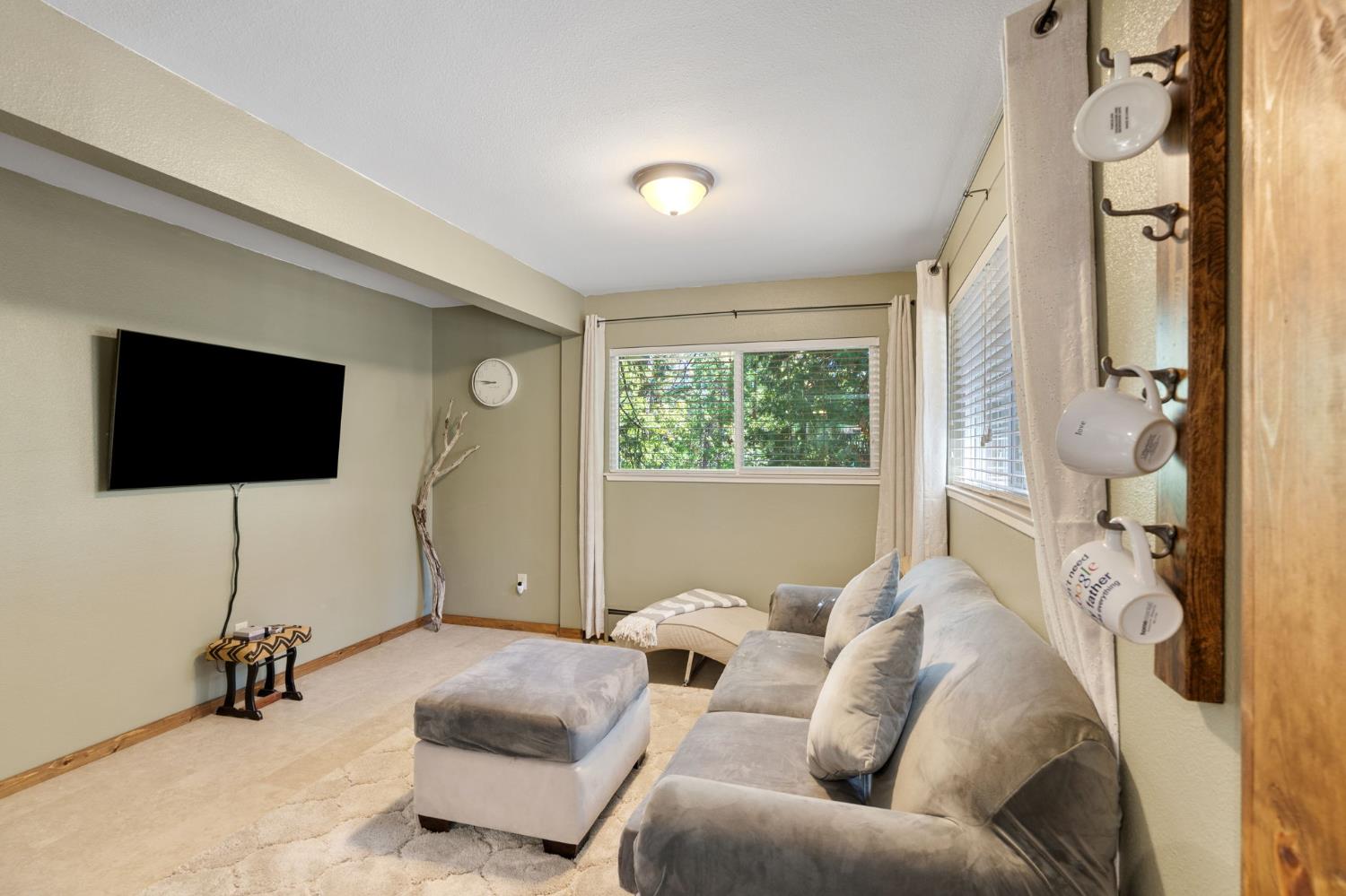 Living room with a gray sectional, ottoman, wall-mounted television, and leafy views through the windows.