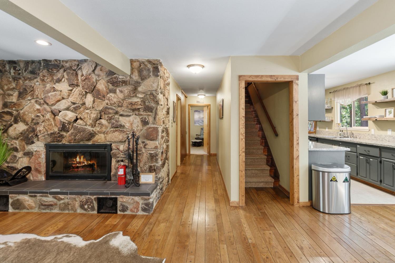 Living area with a stone fireplace, wood floors, and a staircase beside the kitchen.