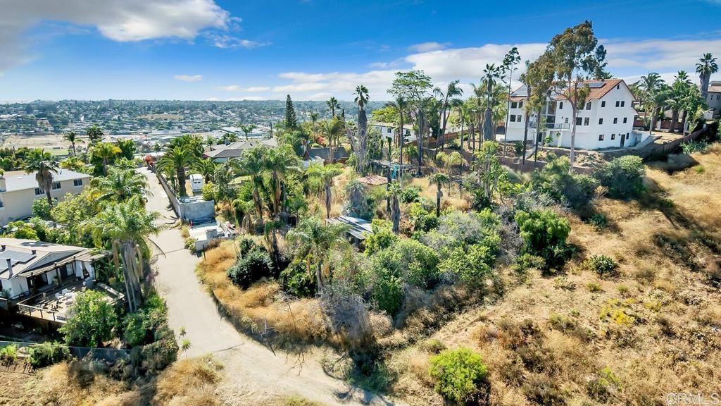 Vacant hillside lot with dry grass, palm trees, nearby homes, and a distant city view.