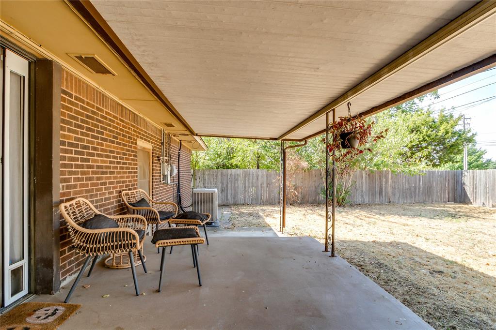 Covered patio with wicker seating beside a brick house, overlooking a fenced backyard.
