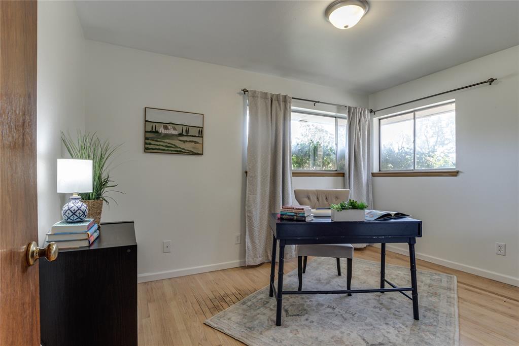 Staged home office with a dark desk, two curtained windows, and hardwood floors.
