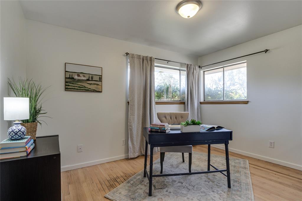 Staged home office with a dark desk and chair between two curtained windows, on hardwood floors.