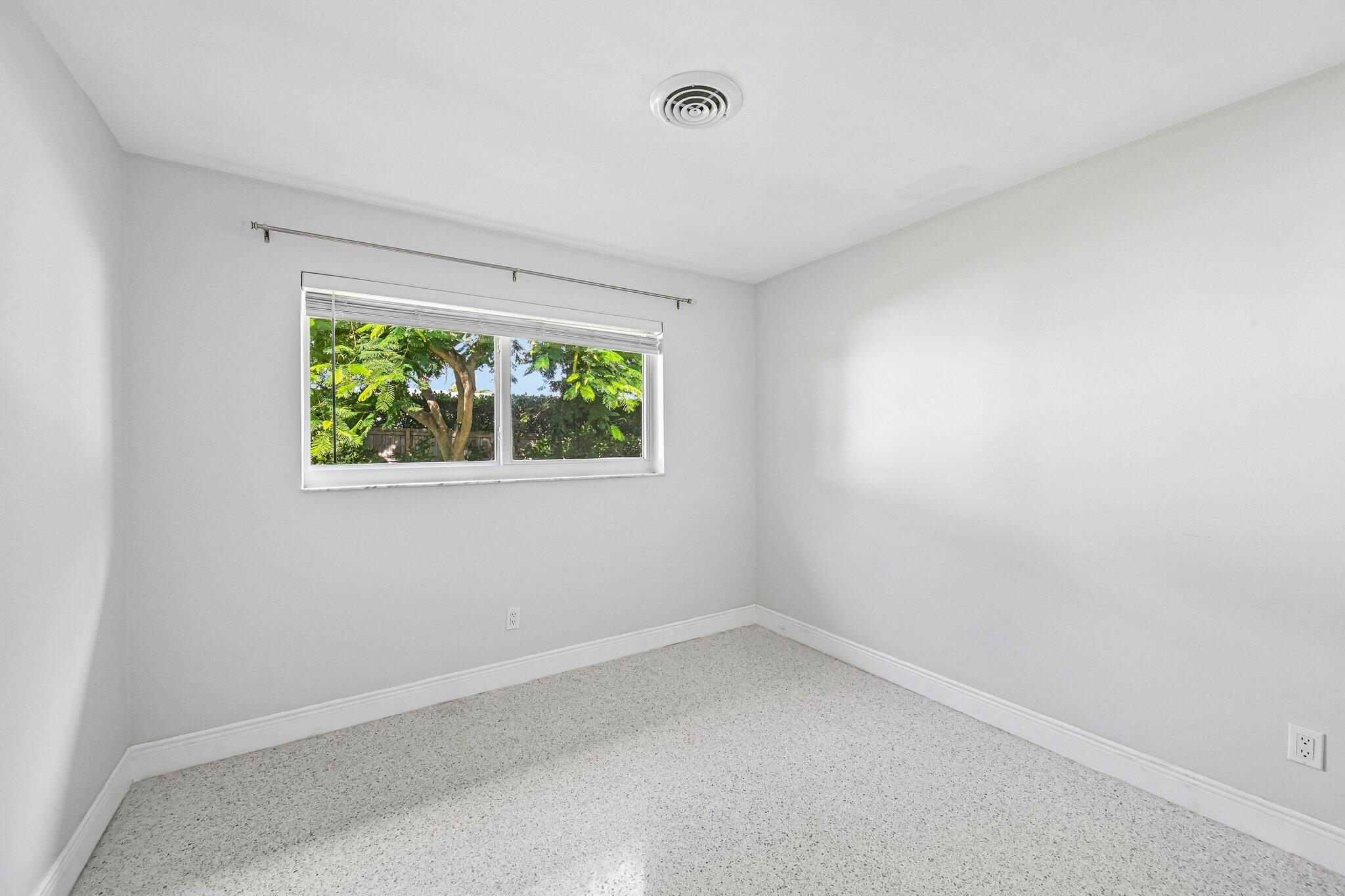 Empty carpeted room with white walls and a window overlooking leafy trees.