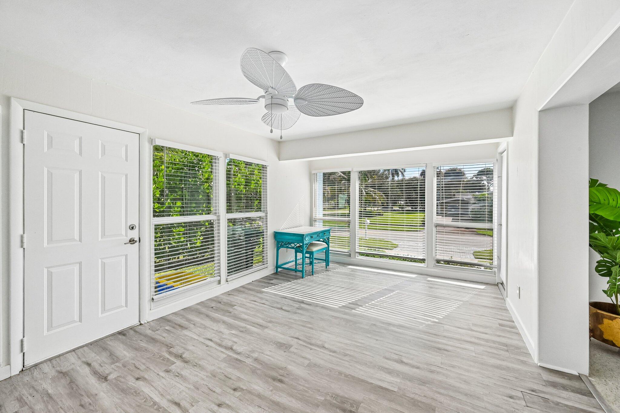 Bright sunroom with wraparound windows, gray plank flooring, and a turquoise table beneath a ceiling fan.