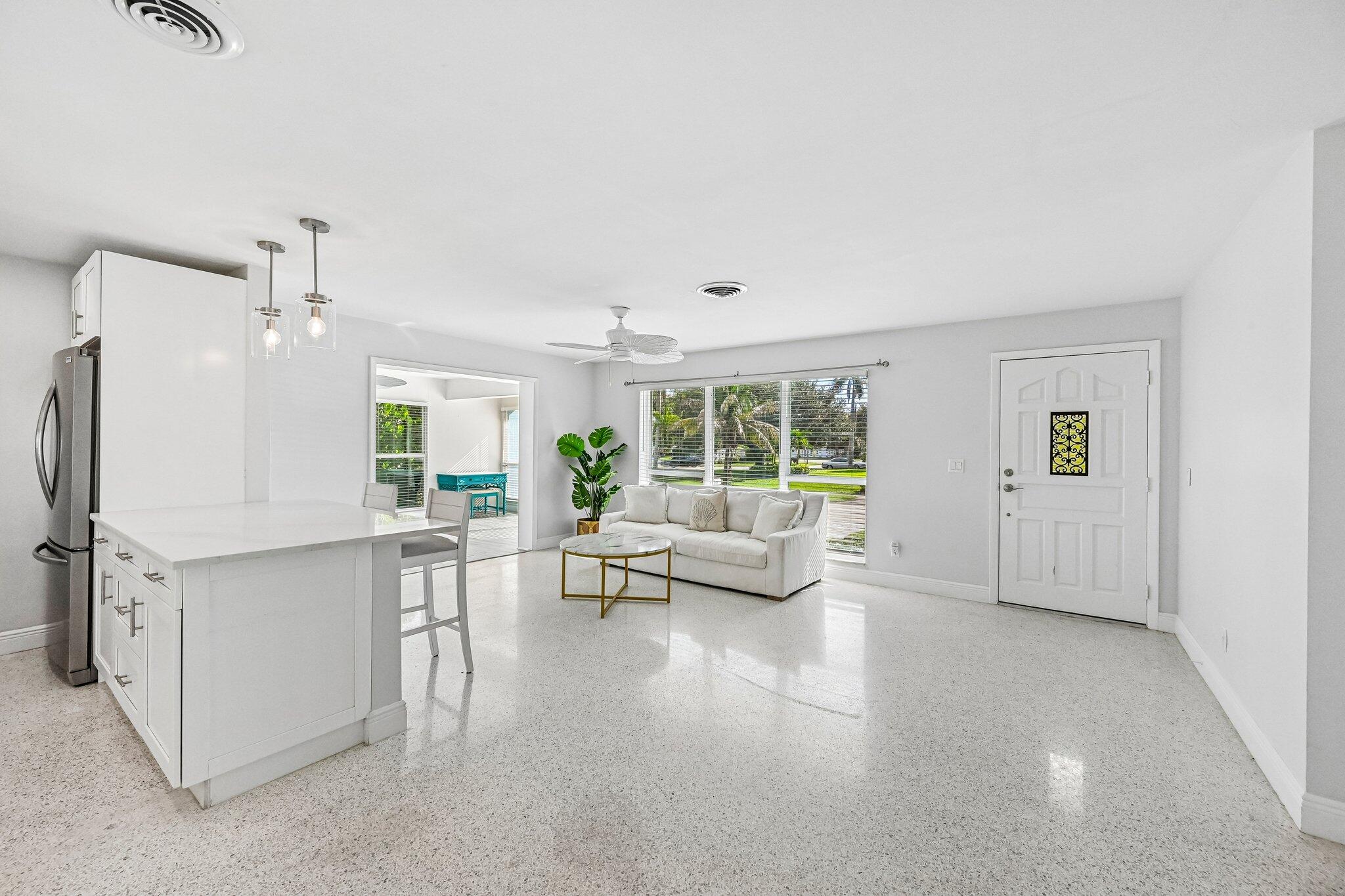 Bright open living room and kitchen with terrazzo flooring, white furnishings, and large garden-facing windows.