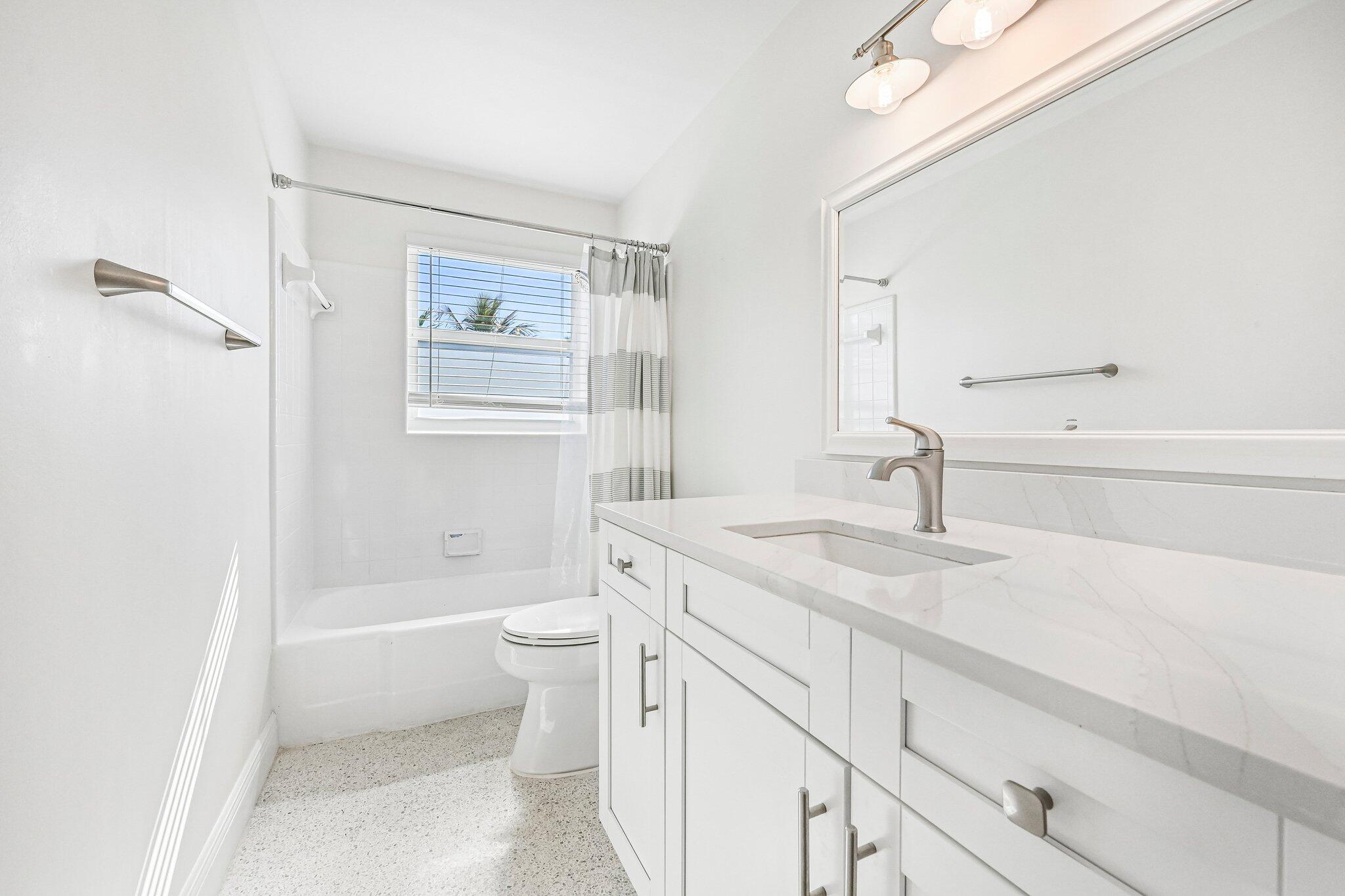 Bright white bathroom with a marble-look vanity, bathtub, and window with blinds.