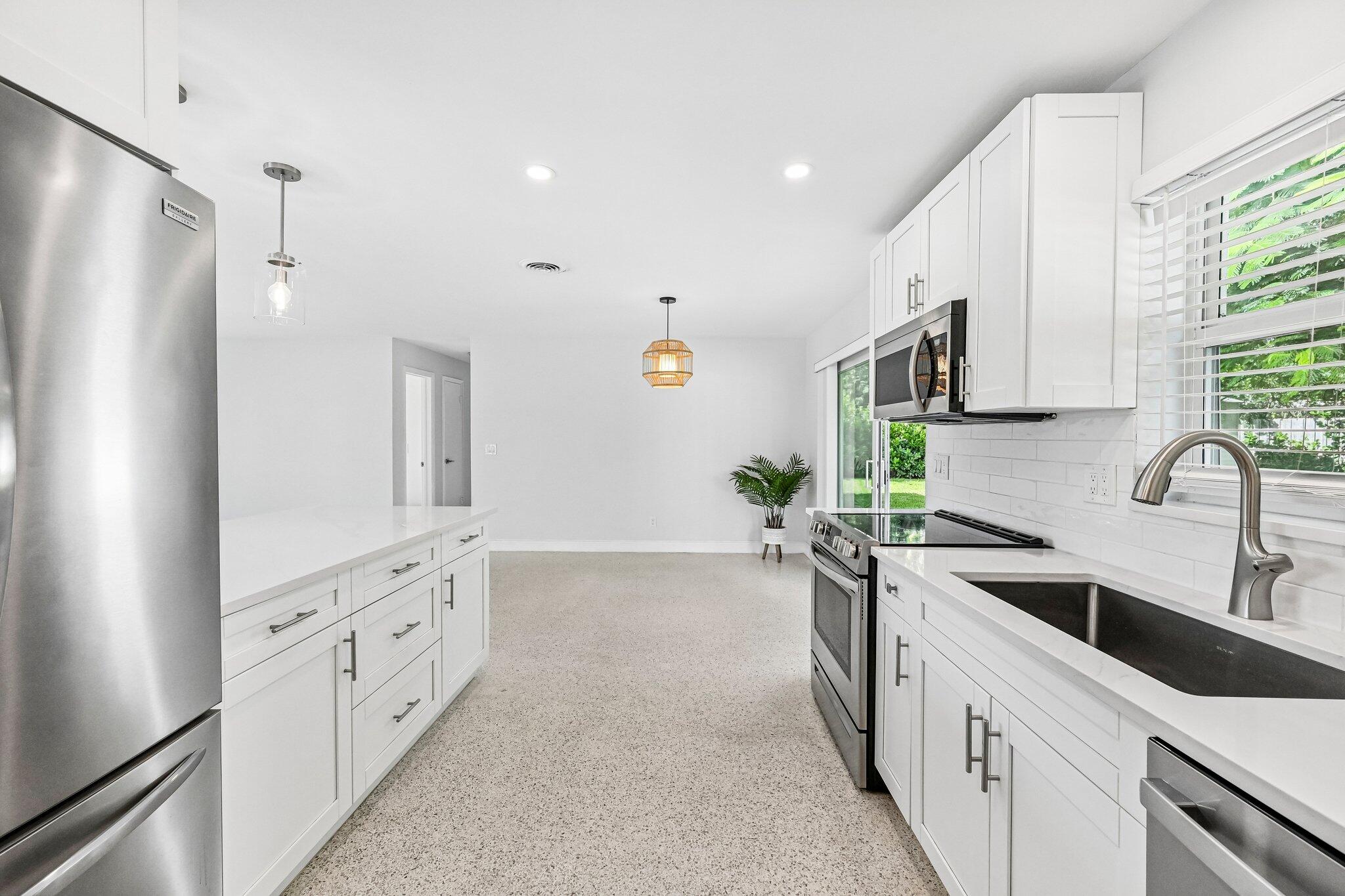 Bright white kitchen with stainless steel appliances, a large island, and a view of greenery through the windows.