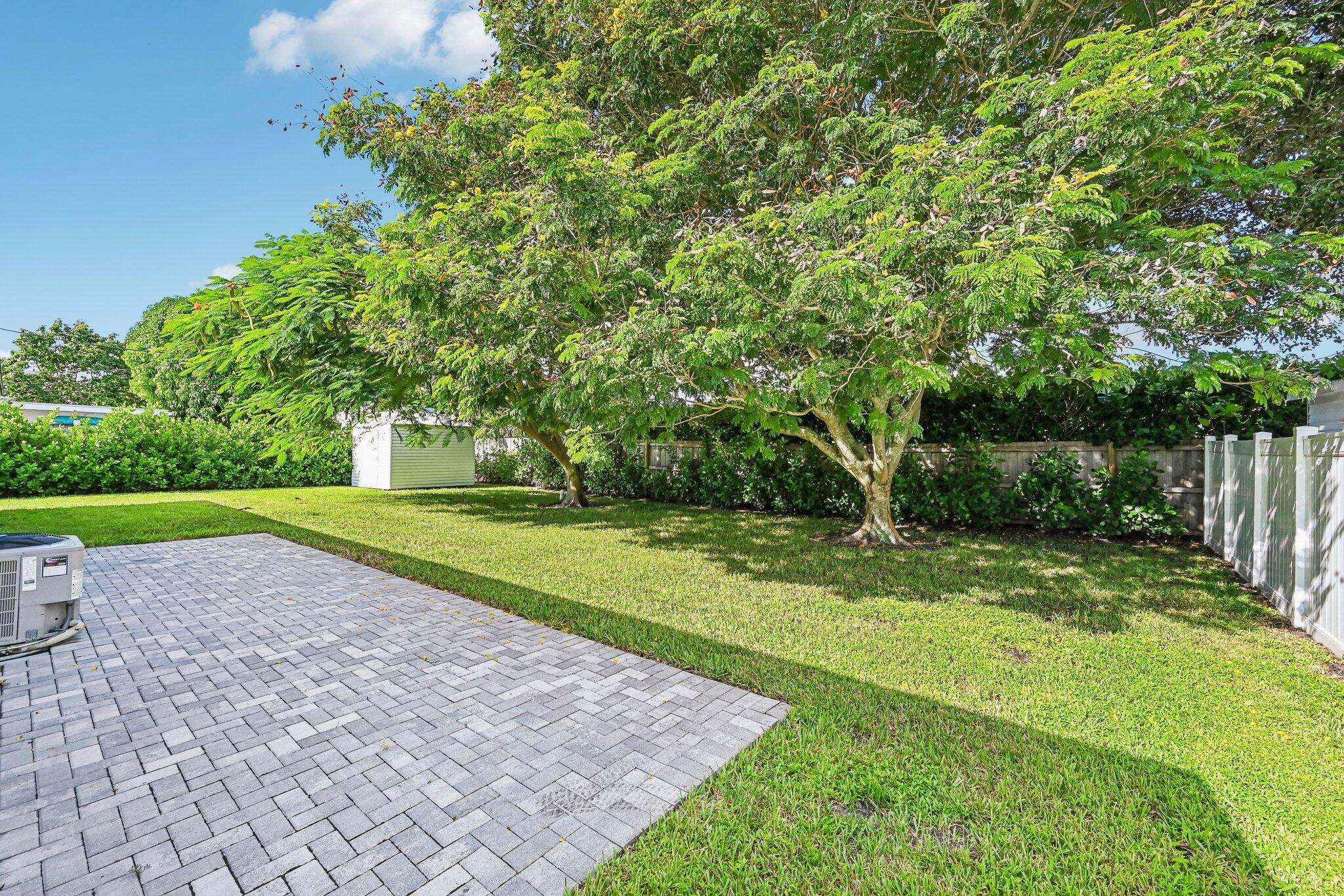 Fenced backyard with a paver patio, broad lawn, mature shade trees, and storage shed.