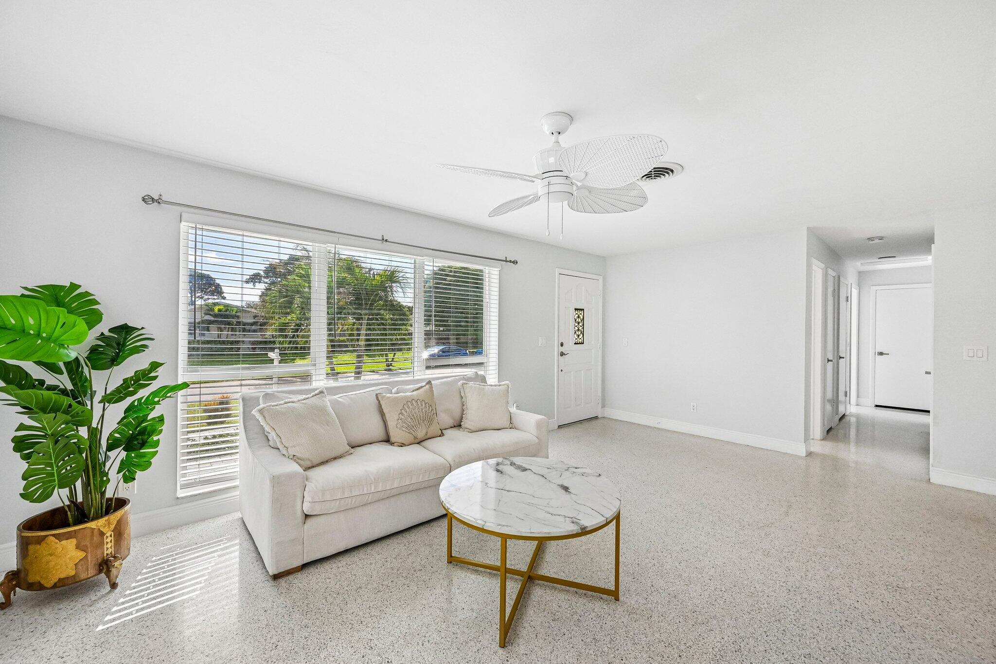 Bright living room with a white sofa, marble-look coffee table, ceiling fan, and large window with blinds.