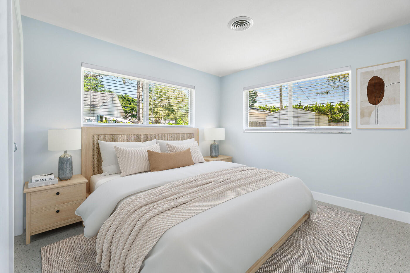 Bright staged bedroom with a woven-headboard bed, pale blue walls, and two wide windows.