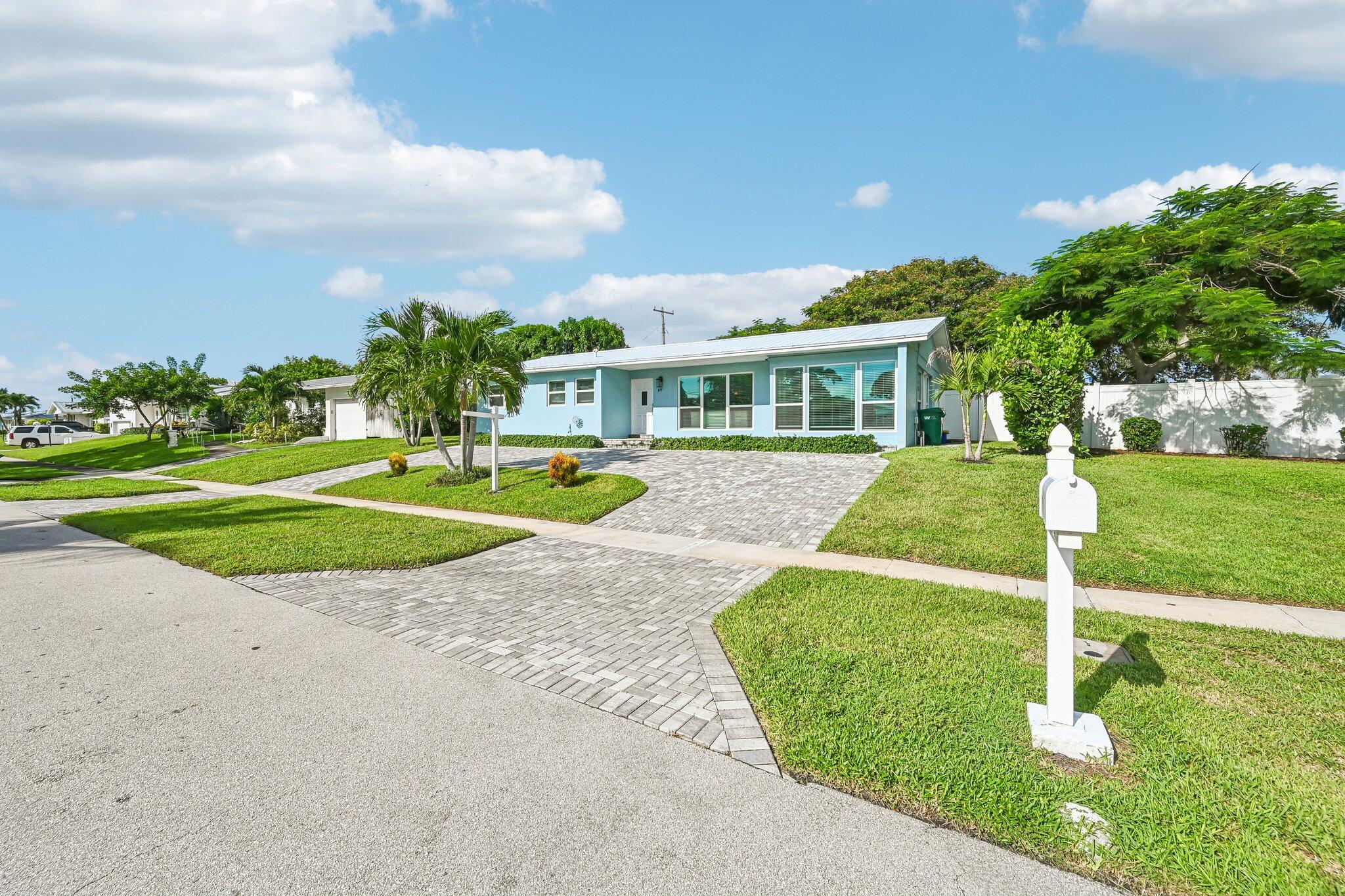 Light blue single-story home with a wide paver driveway and palm trees.