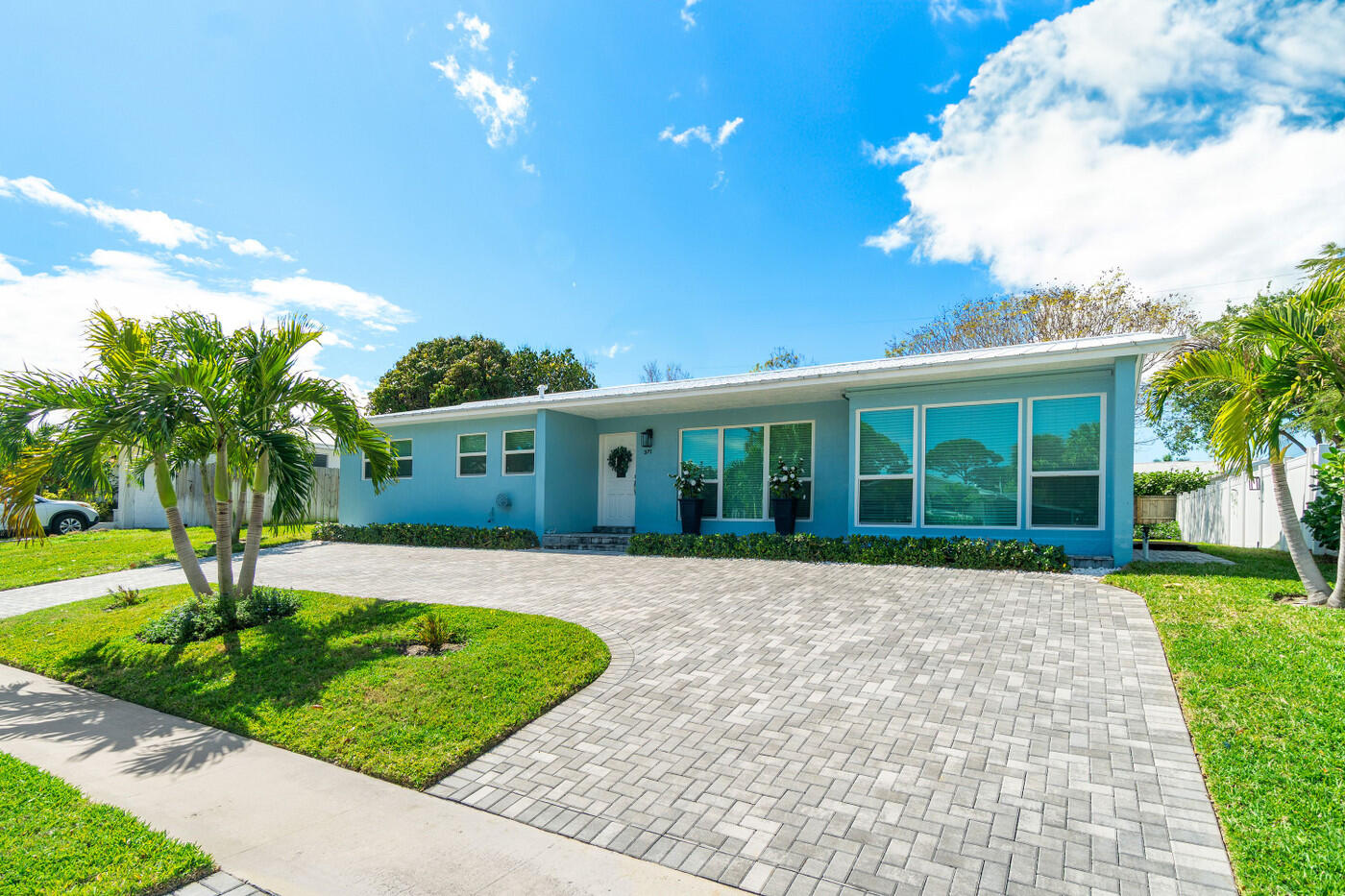 Blue single-story home with large front windows, palm trees, and a curved paver driveway.