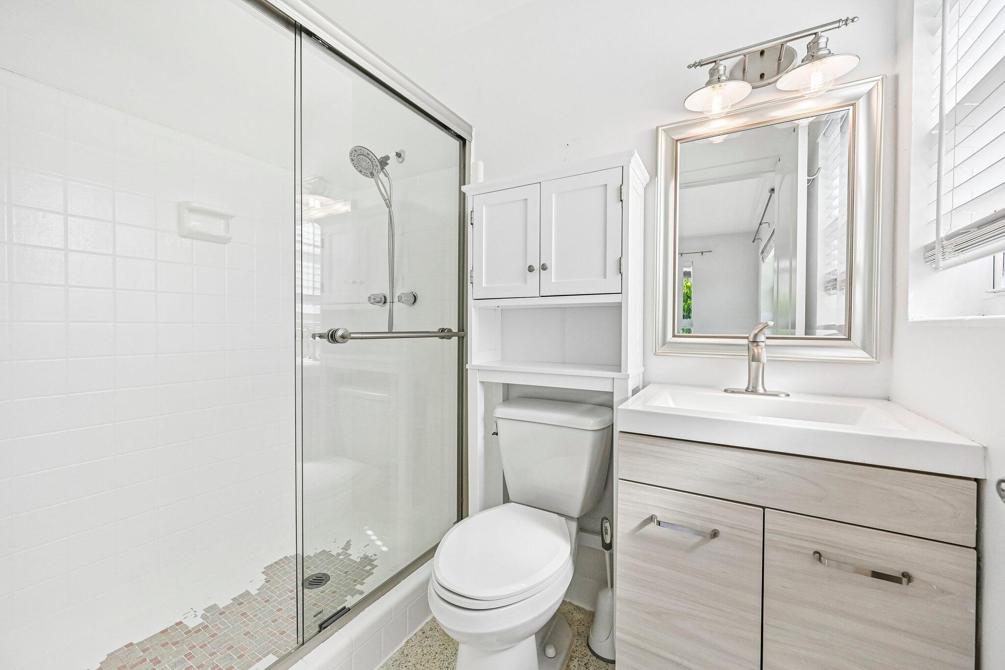 Bright white bathroom with a glass-enclosed shower, toilet, and light wood vanity.