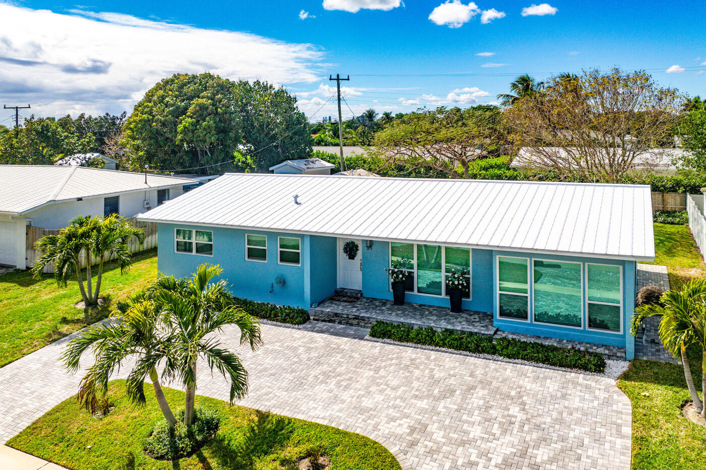 Blue single-story home with a white metal roof and palm-lined paver driveway.