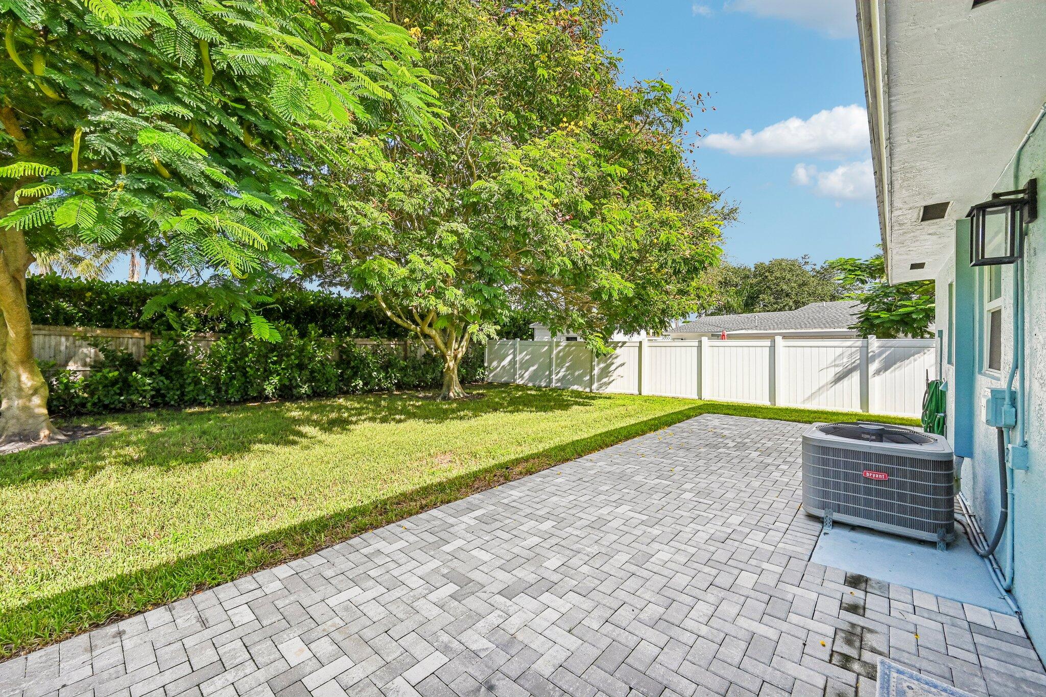Paver patio beside a blue home, overlooking a fenced lawn shaded by mature trees.