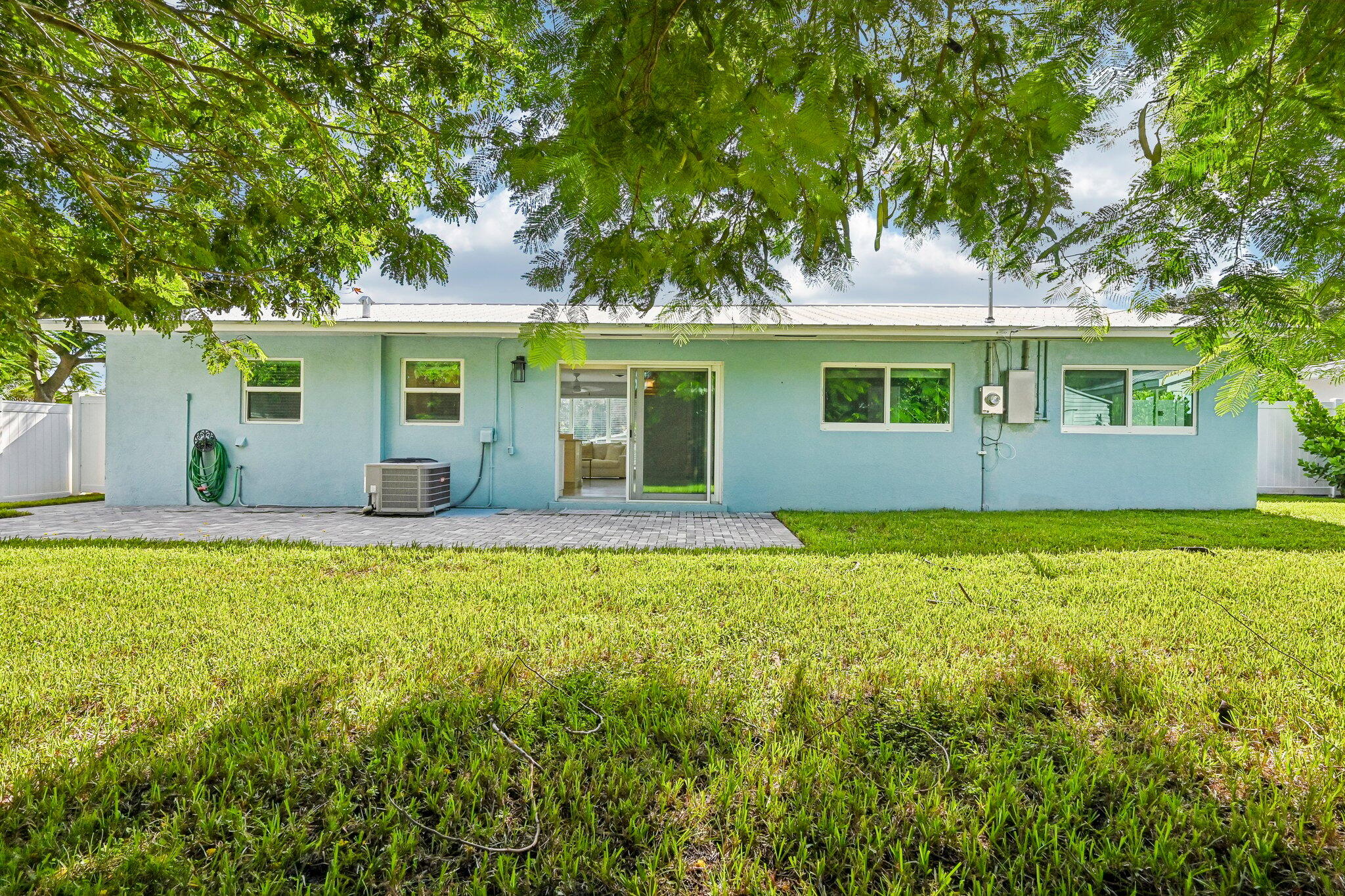 Rear exterior of a light-blue single-story home beneath mature trees, with a paved patio and broad lawn.