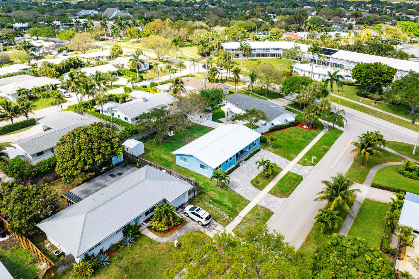 Aerial view of a blue single-story home with a white roof and stone driveway among palm-lined streets.