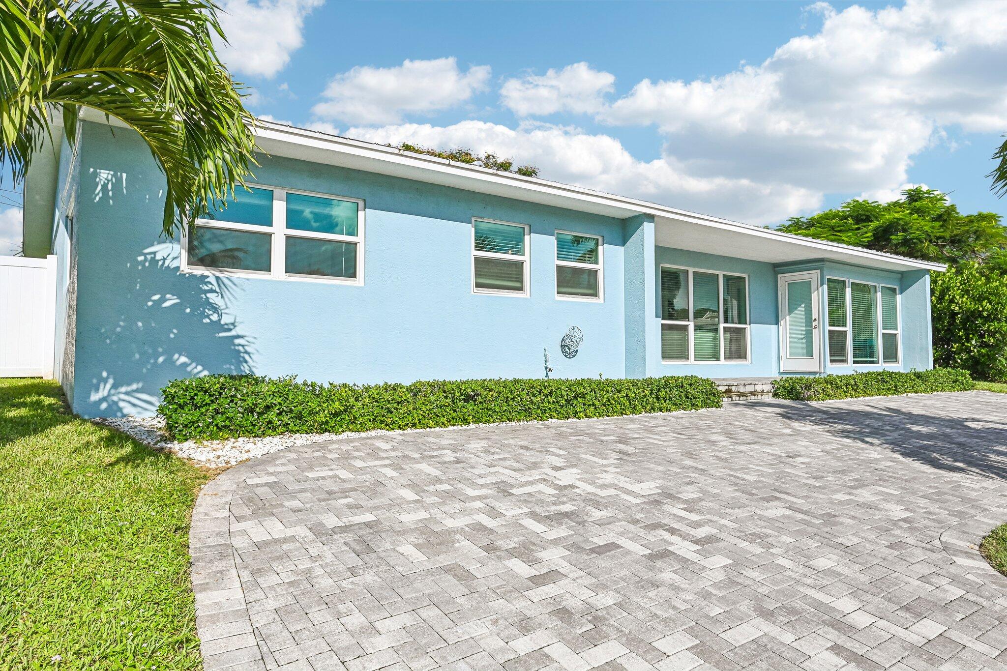 Light blue single-story home with a wide gray paver driveway and tropical landscaping.