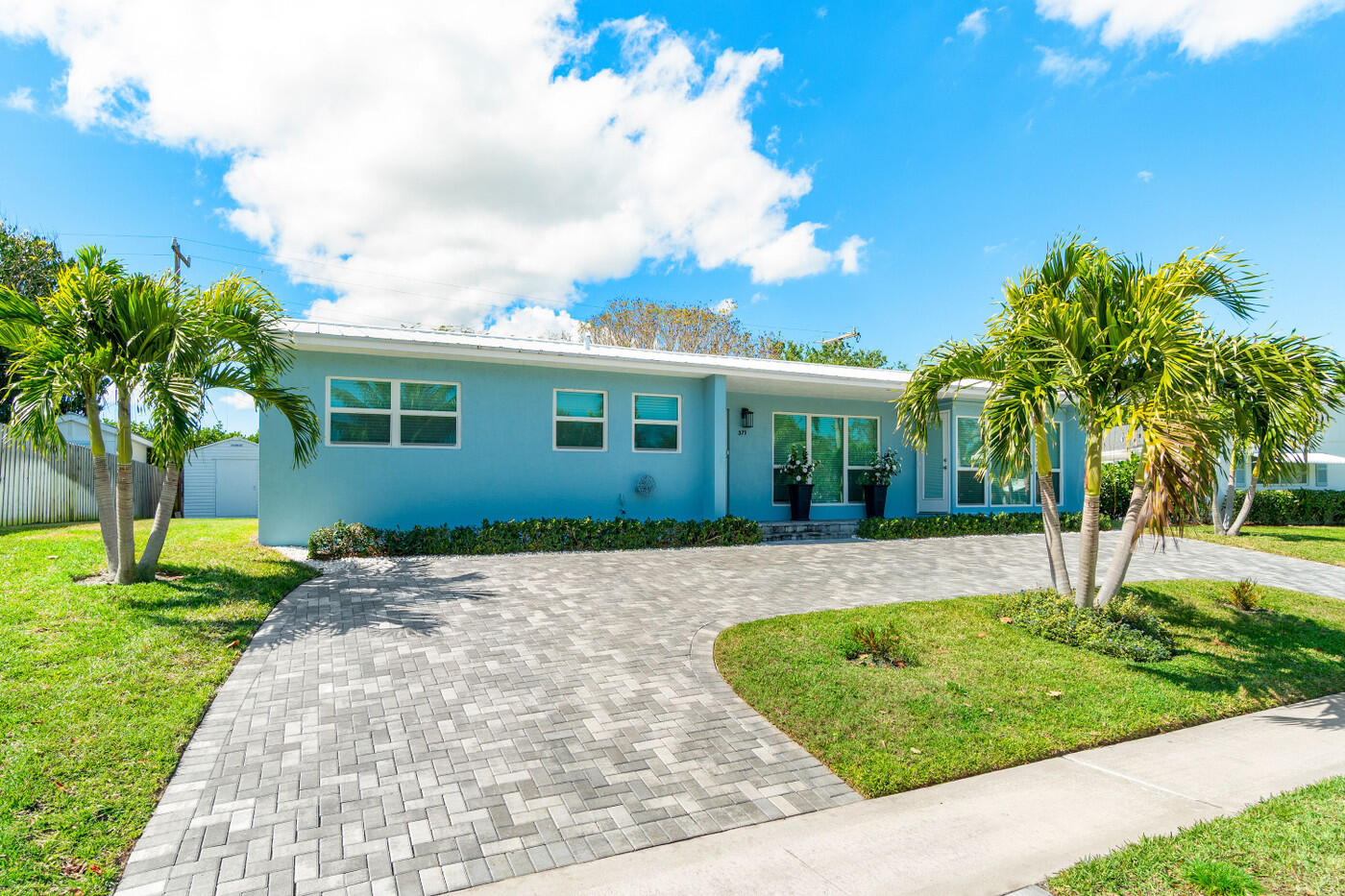 Blue single-story home with a curved paver driveway and palm trees.