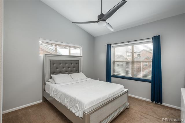 Bedroom with a white bed, tufted gray headboard, ceiling fan, and blue curtains framing a large window.
