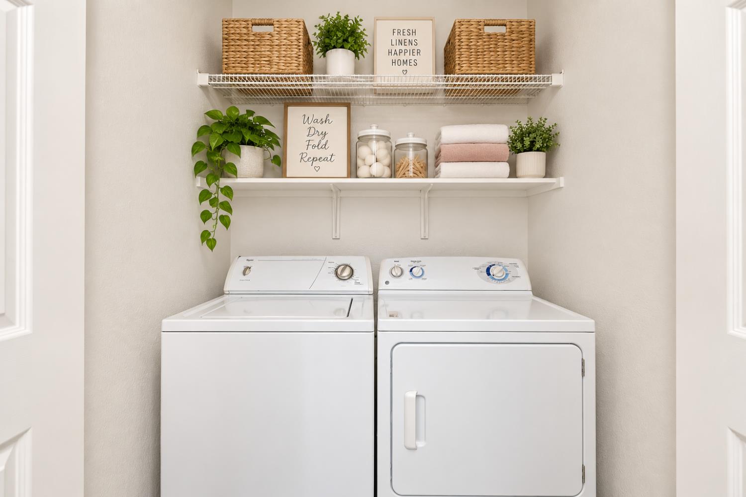 Laundry room with white washer and dryer beneath shelves of baskets, folded towels, and plants.