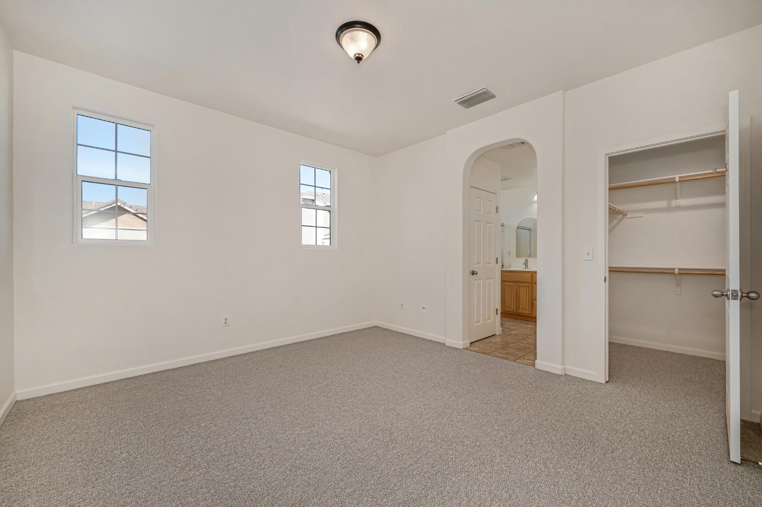 Empty carpeted bedroom with two windows, an open closet, and an arched bathroom doorway.
