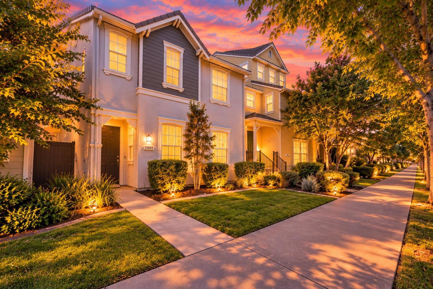 Two-story home at dusk with illuminated windows, landscaped frontage, and a tree-lined sidewalk.