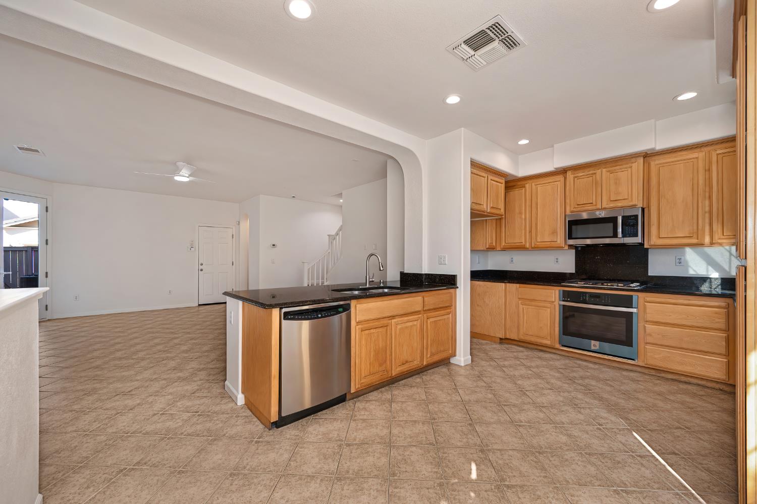 Kitchen with wood cabinets, dark counters, stainless steel appliances, and an island opening to a living area.