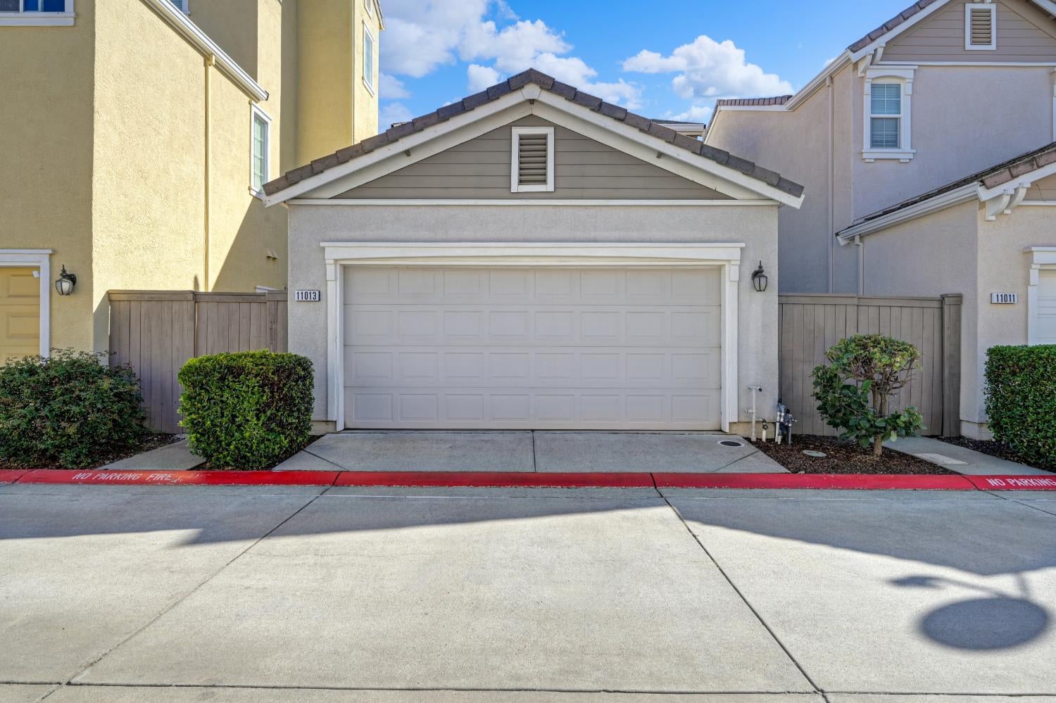 Gray stucco home with a double garage, gabled roof, and shrubs beside the driveway.