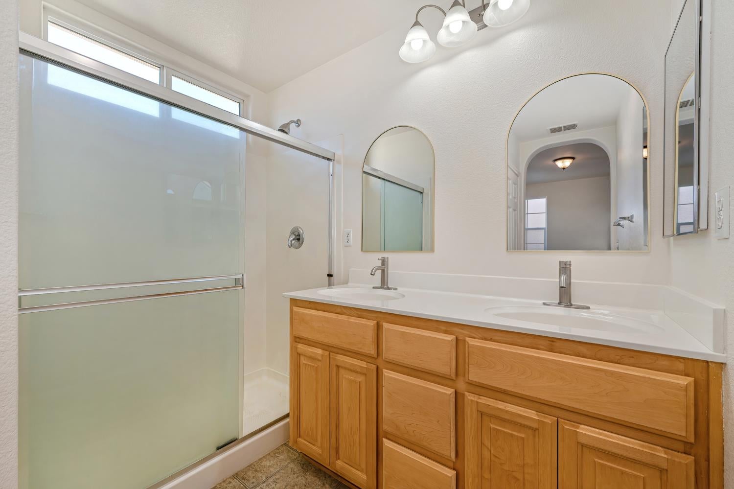 Bathroom with a double-sink wood vanity, arched mirrors, and frosted-glass shower doors.