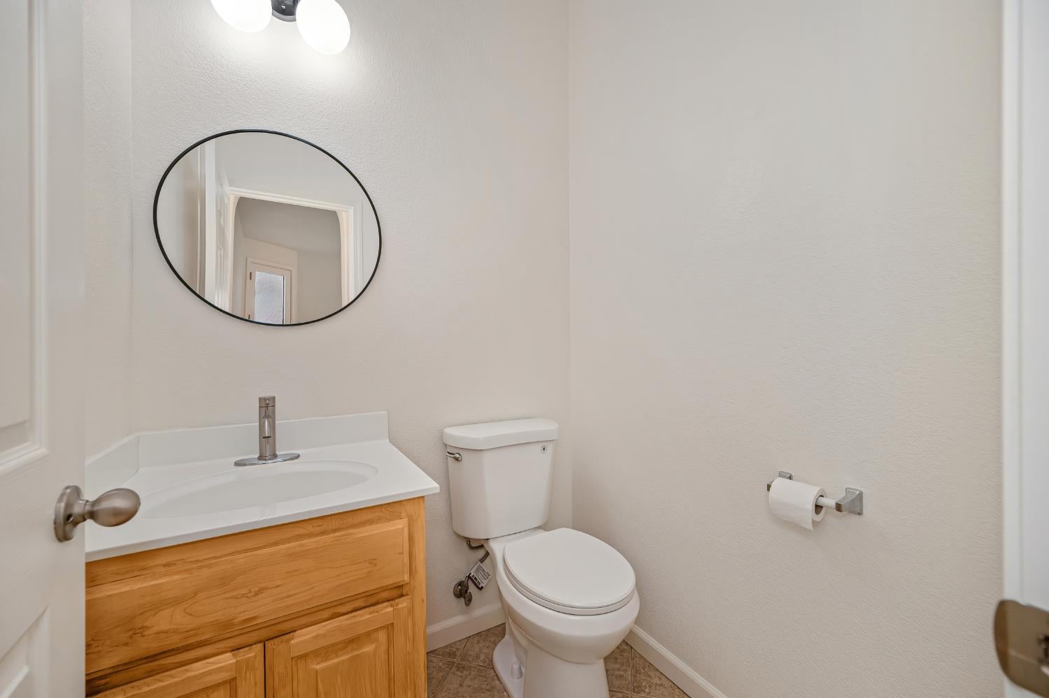 Small bathroom with a wood vanity, white sink, toilet, and oval mirror.
