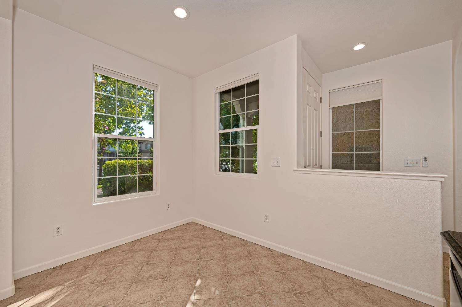 Empty kitchen nook with beige tile flooring and windows overlooking greenery.