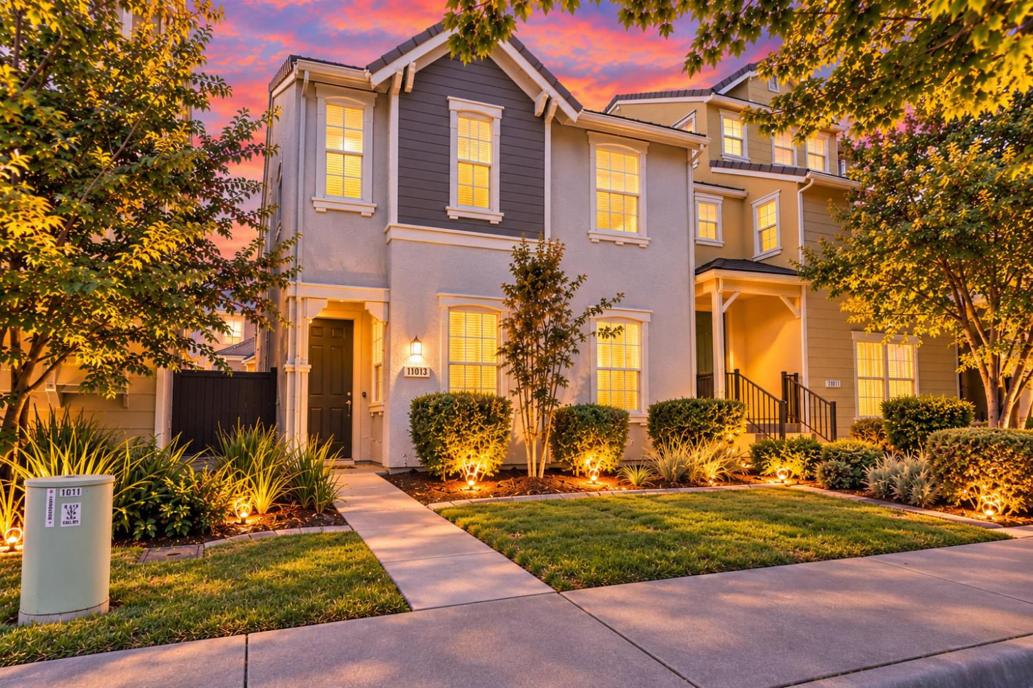 Two-story home with illuminated windows, landscaped front yard, and a vivid sunset sky.