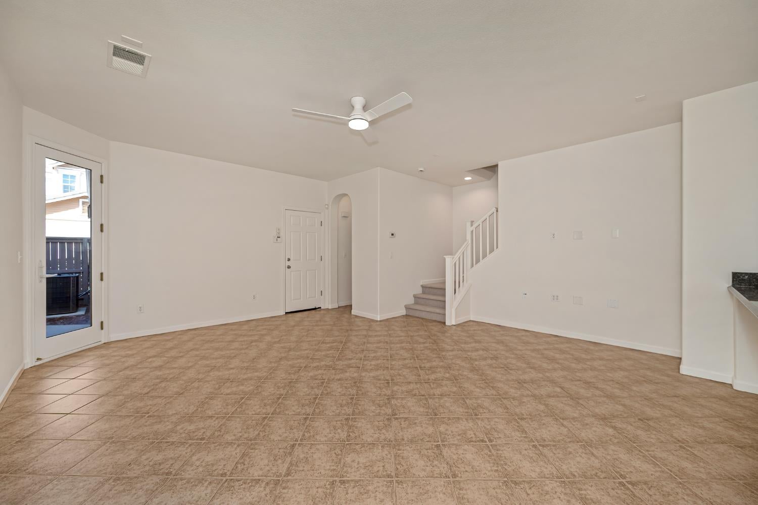 Empty living area with tan tile floors, a ceiling fan, and a carpeted staircase.